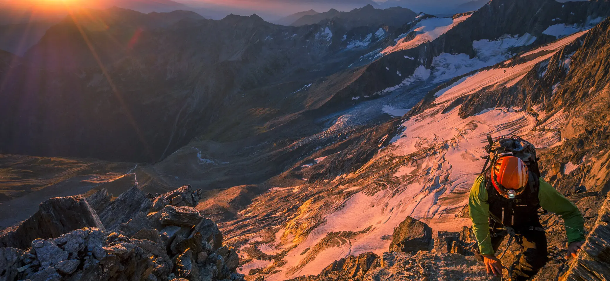 Bergsteiger mit Stirnlampe vor der untergehenden Sonne, umgeben von Bergen, teilweise mit Schneefeldern im Hintergrund. | © DAV/Silvan Metz